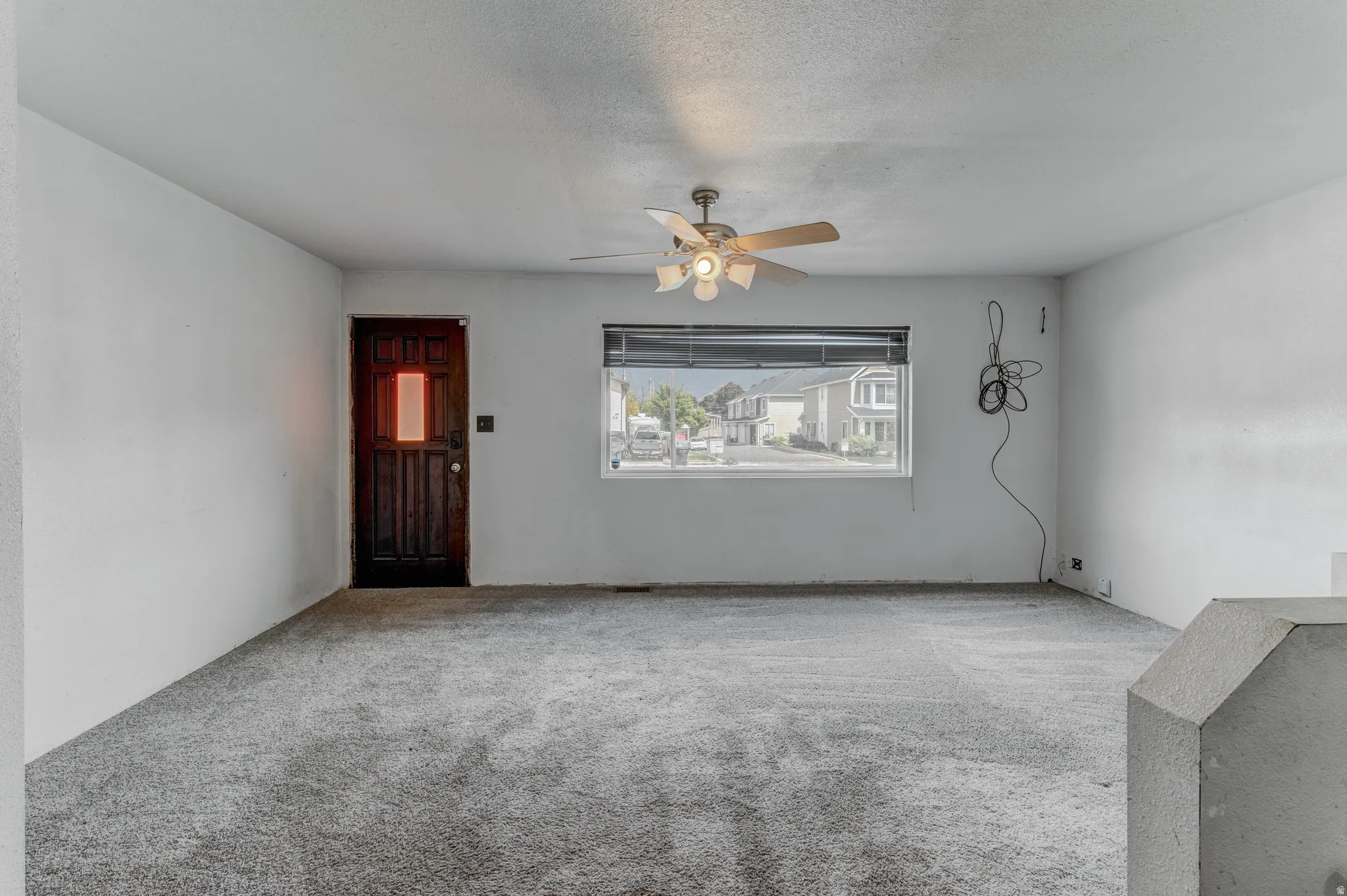 Spacious room with light gray carpeting, a dark wood-paneled door with a rectangular window, and a window with a neighborhood backdrop
