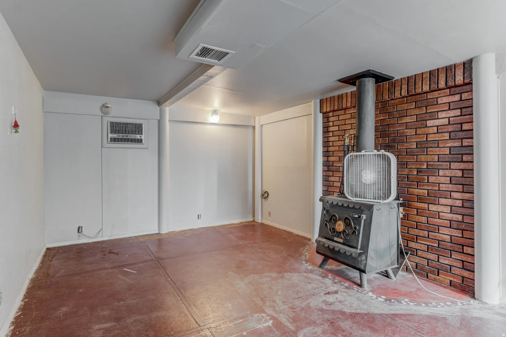 Exposed brick accent wall, wood-burning stove with metal flue, concrete flooring, and an in-wall air conditioning unit