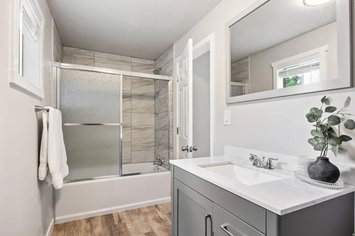 Wood-finish flooring, full bathtub with sliding glass doors, tiled shower surround, framed mirror, and a vanity with a white countertop and integrated sink