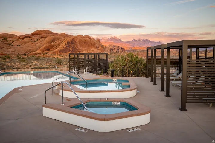 Outdoor aquatic area featuring multiple pools, two integrated spas with handrails, a paved deck, and a series of cabanas with horizontal slatted walls