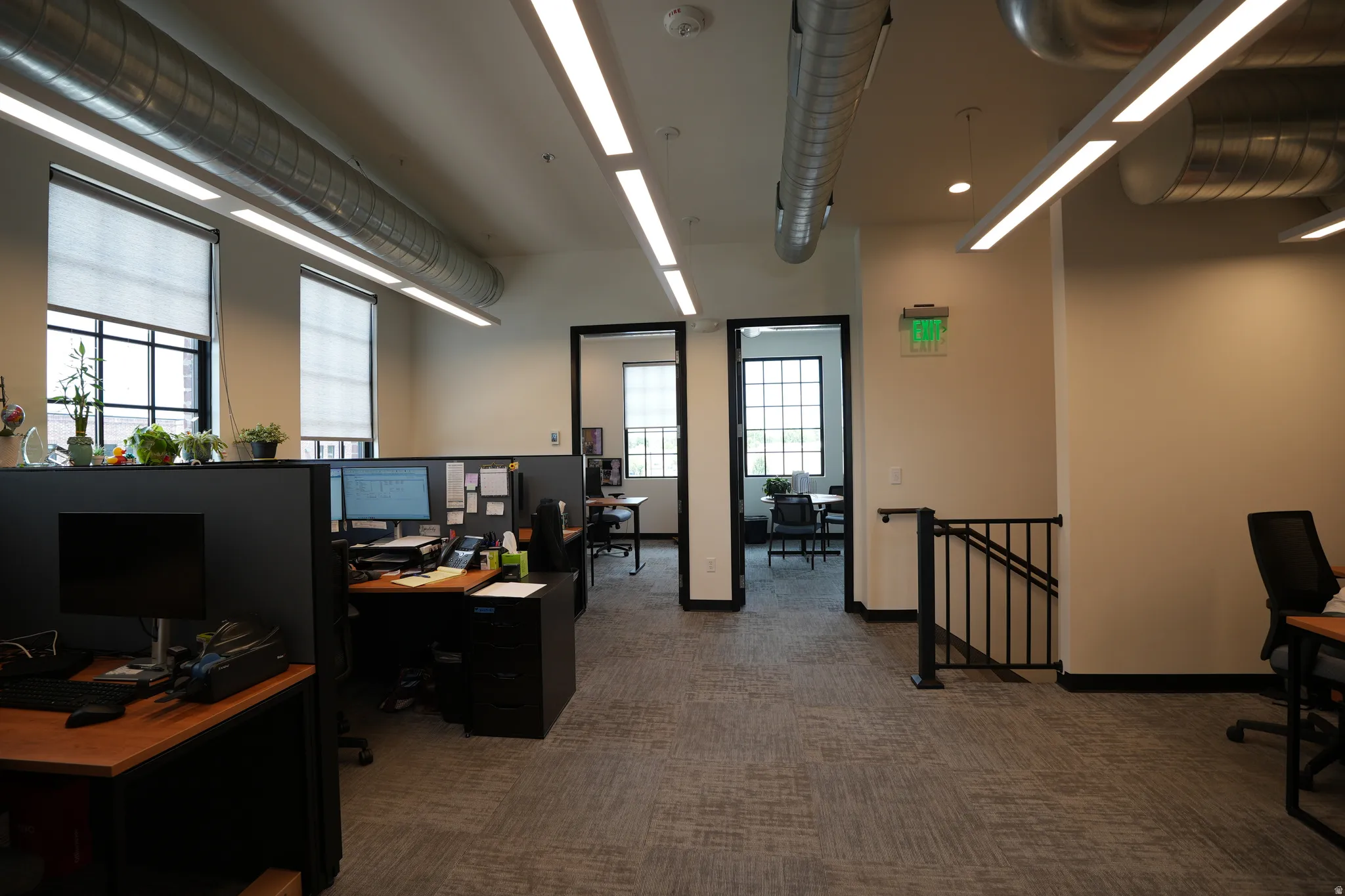 Open-concept office space featuring carpet tile flooring, exposed spiral ductwork, and abundant natural light from large windows