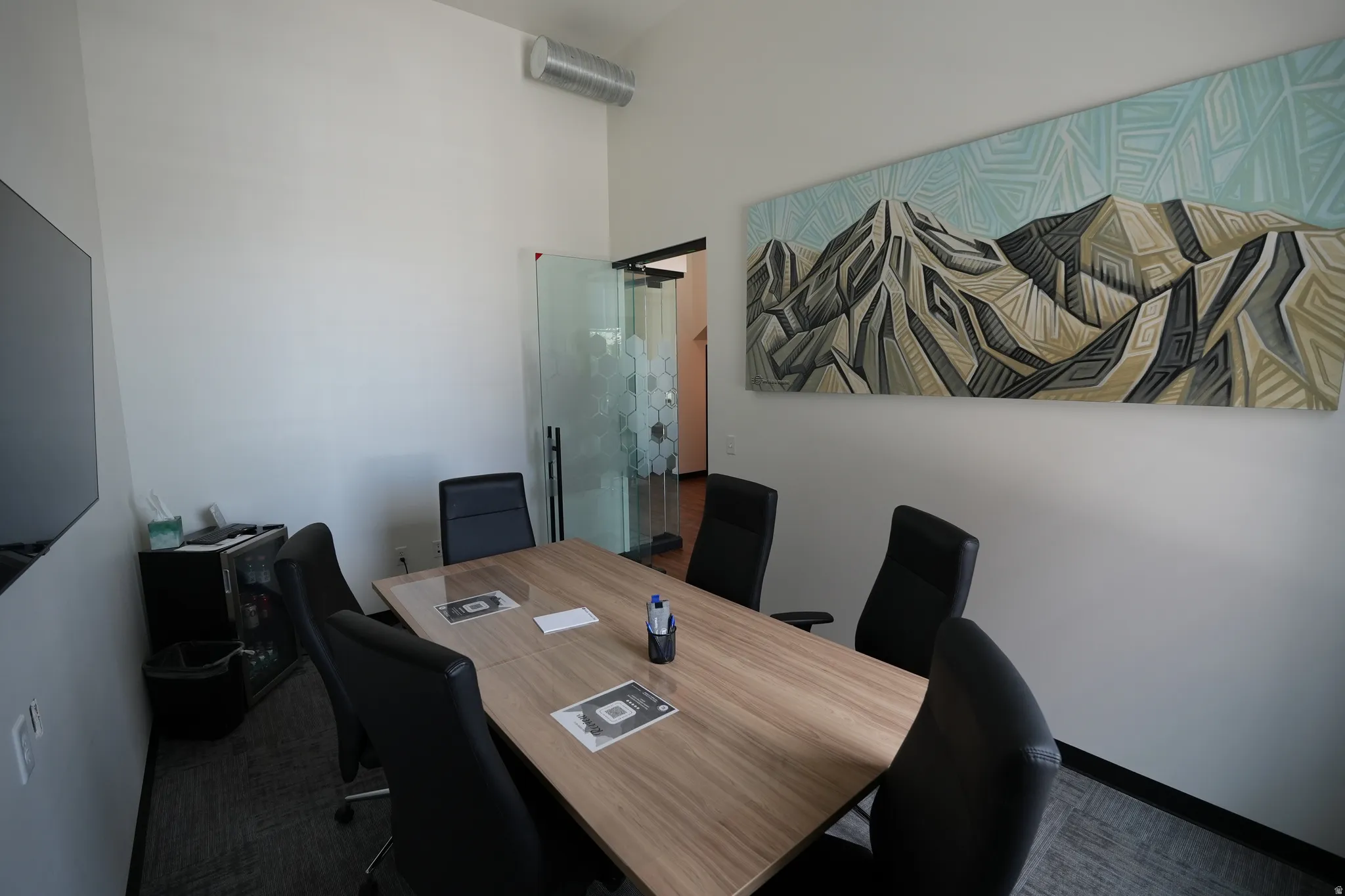 Angled ceiling with exposed ductwork, frosted glass partition with geometric pattern, wood-finish conference table, and textured carpet flooring