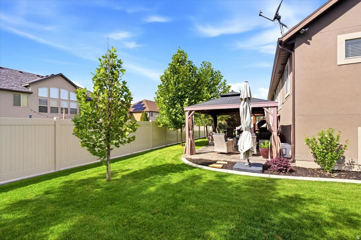 Expansive lawn area with a privacy fence, established trees, and a paved patio featuring a built-in gazebo
