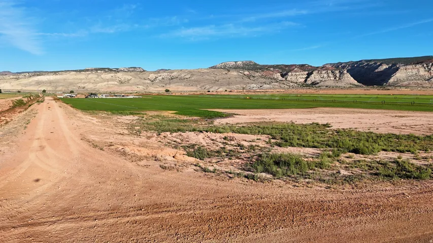 Expansive land featuring a dirt road, distant structures, and lush green fields, all set against a backdrop of mountainous terrain
