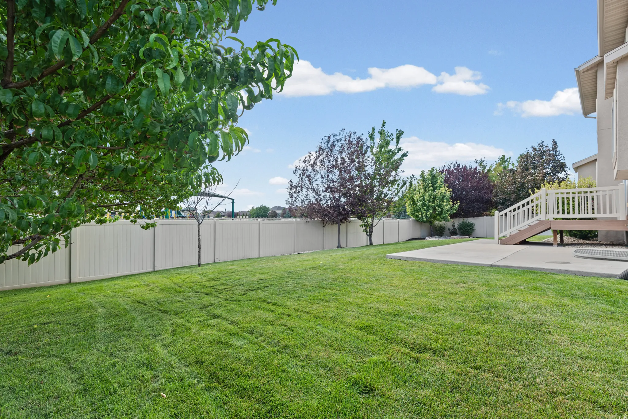 Expansive grass lawn with mature trees, a private perimeter fence, a concrete patio, and an elevated wood deck with stairs