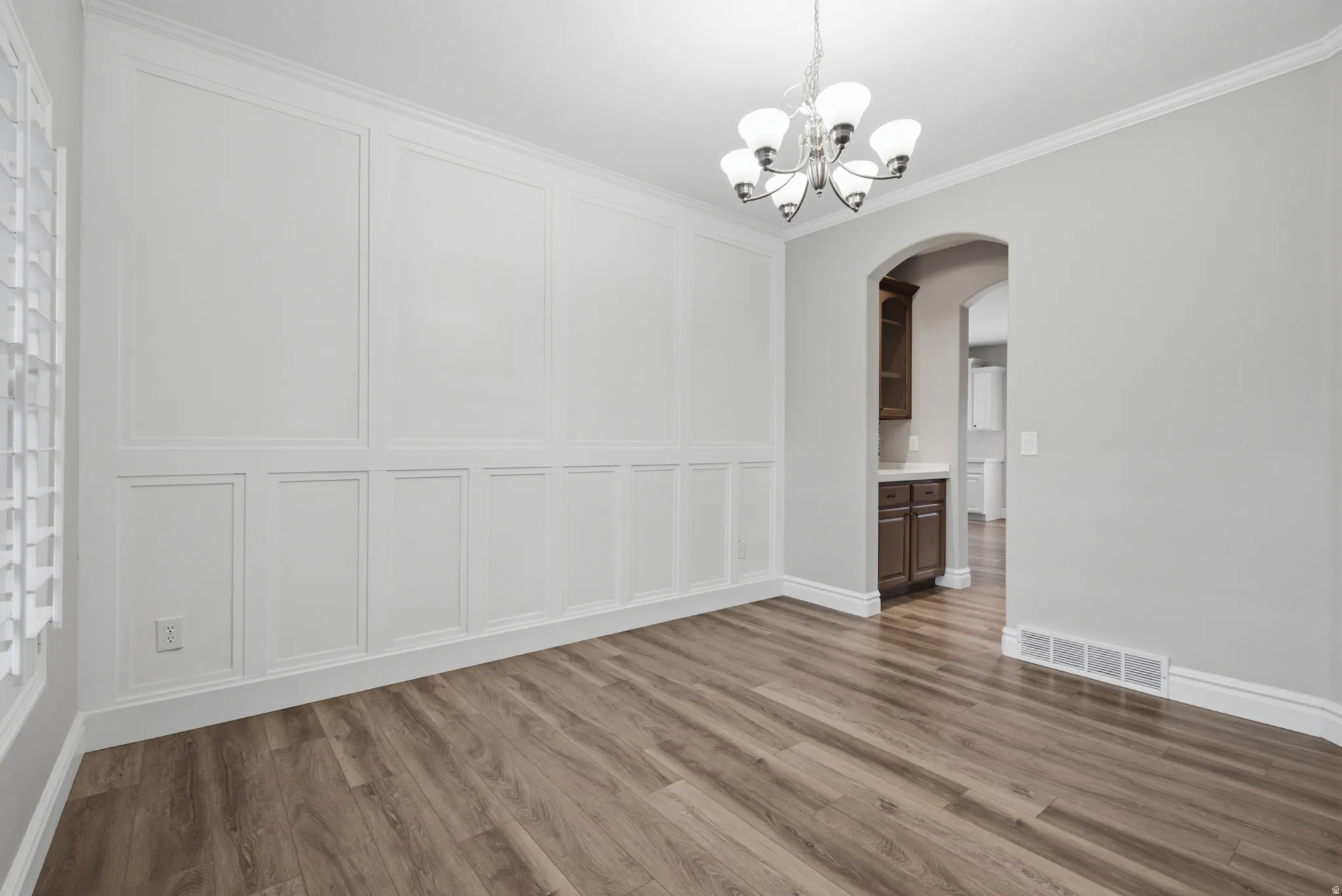 Wood-finish flooring, decorative wall paneling, crown molding, and a chandelier illuminate this versatile space