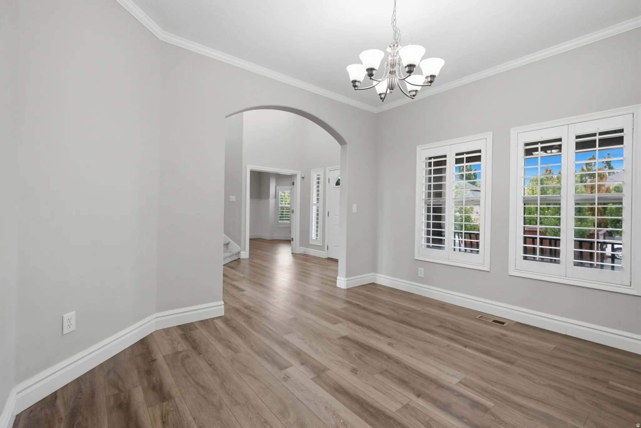 Wood-finish flooring, decorative archway, crown molding, two windows with plantation shutters, and a multi-arm chandelier