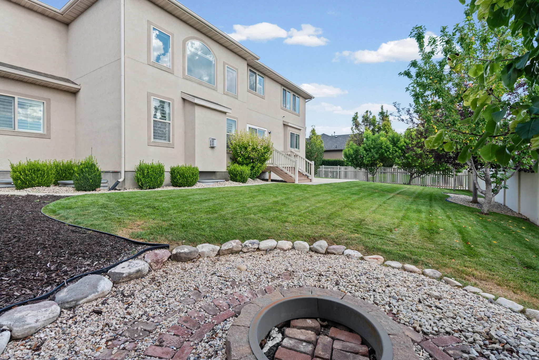 Expansive backyard with a manicured lawn, established trees, and a built-in fire pit with stone and brick accents