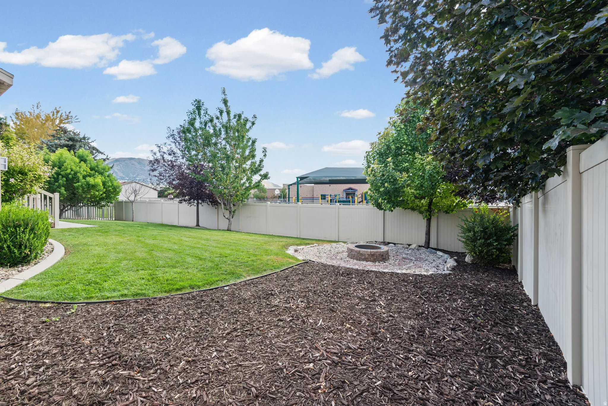 Expansive backyard featuring a lush lawn, mature trees, a stone-ringed fire pit, decorative rock landscaping, and a privacy fence