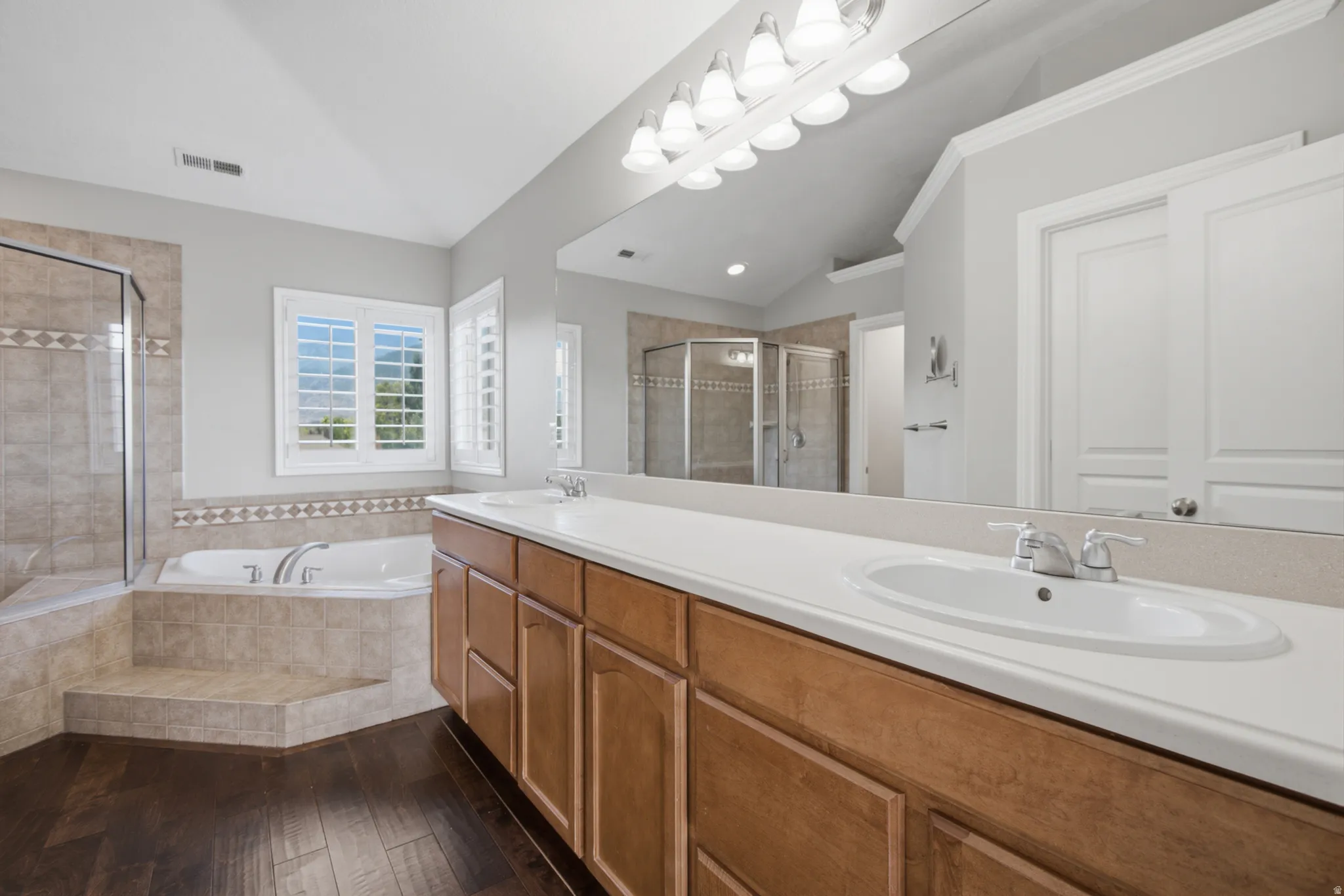 Dual vanity with integrated sinks, wood-finish cabinetry, tile-surround soaking tub, separate glass-enclosed shower, and plantation shutters