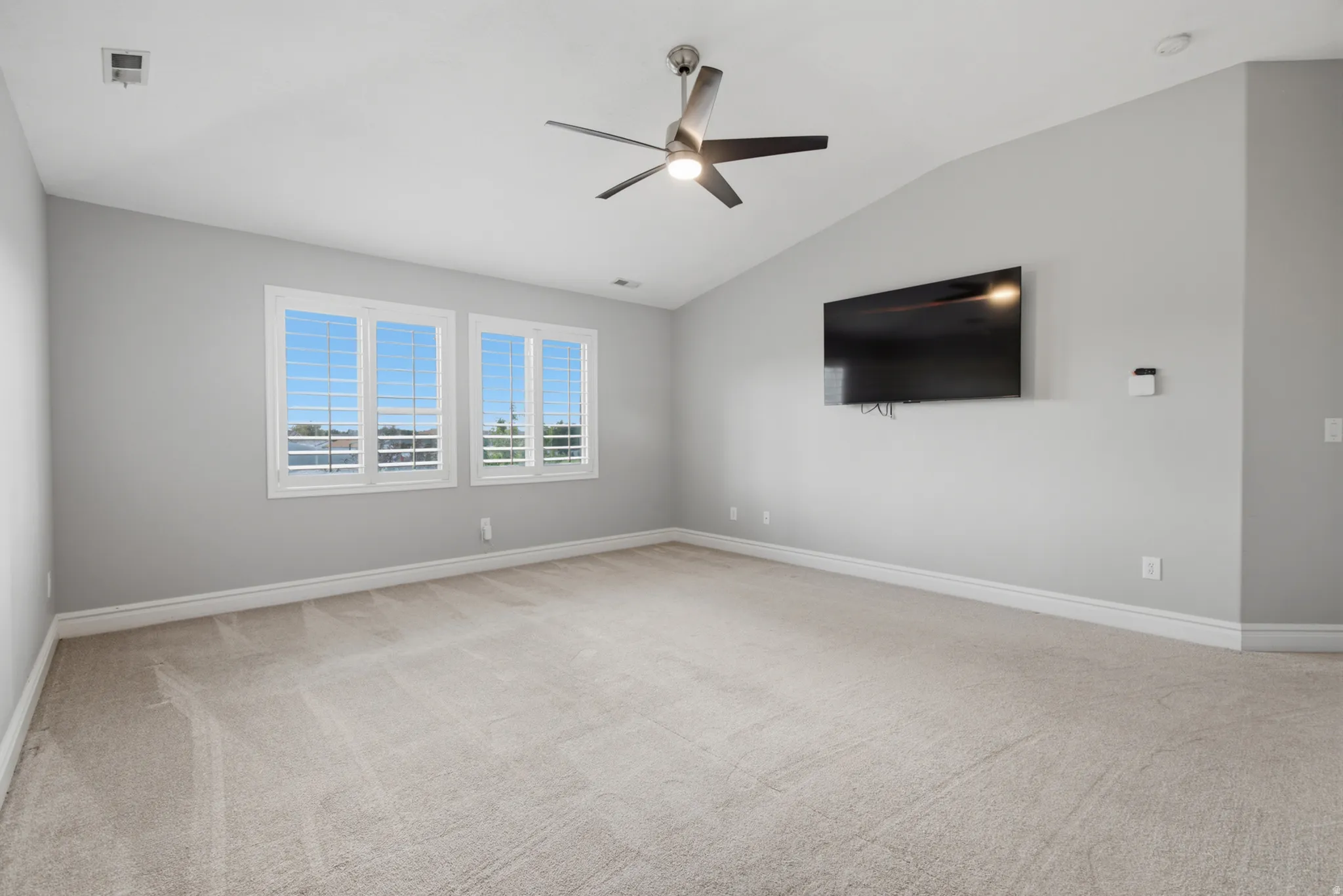 Vaulted ceiling with a ceiling fan, plush carpet flooring, multiple windows with white plantation shutters, and a wall-mounted television