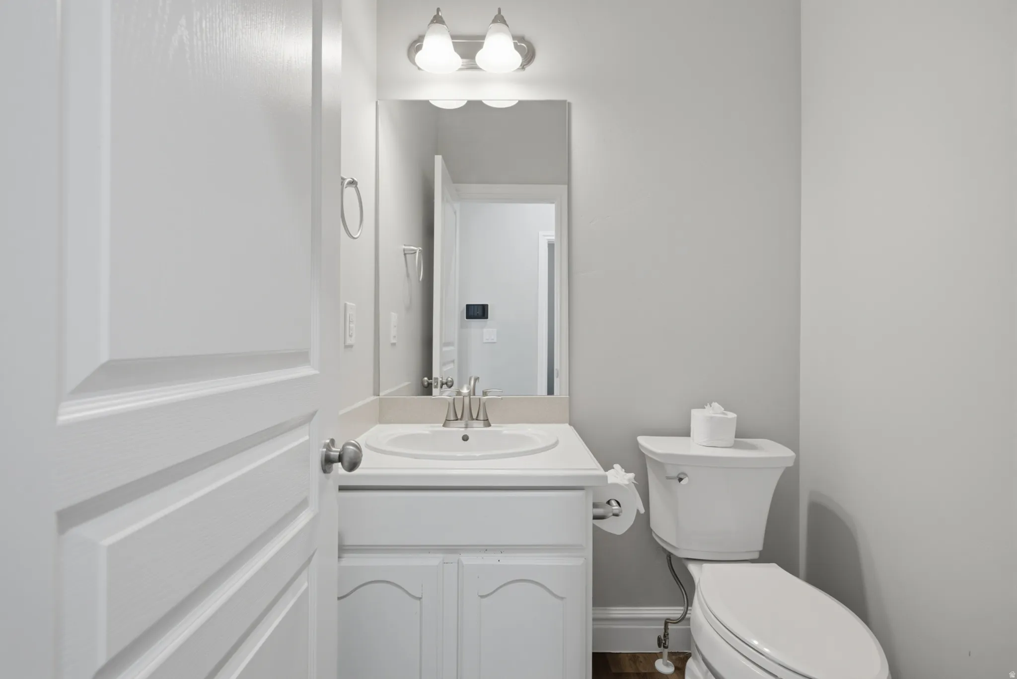 White paneled door opening to a bathroom featuring a vanity with an integrated sink, a rectangular mirror, and a wall-mounted light fixture with two sconces