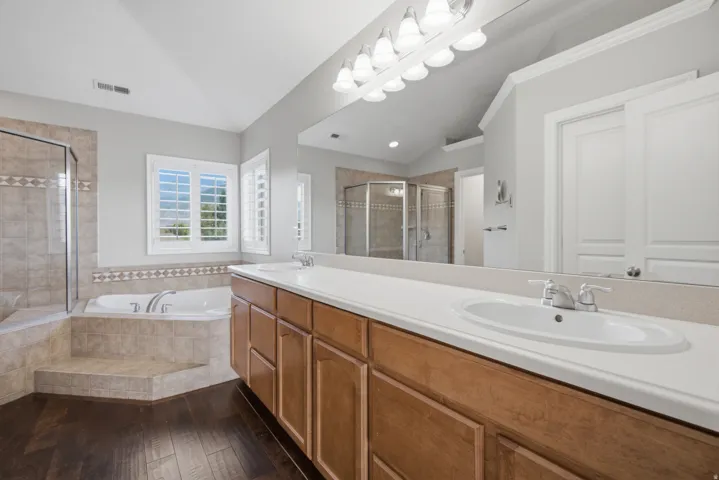Dual vanity with integrated sinks, wood-finish cabinetry, tile-surround soaking tub, separate glass-enclosed shower, and plantation shutters