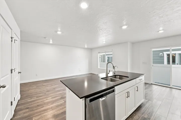Kitchen island with a dark countertop and undermount double sink, stainless steel dishwasher, white cabinetry with dark pulls, wood-finish flooring, and recessed lighting