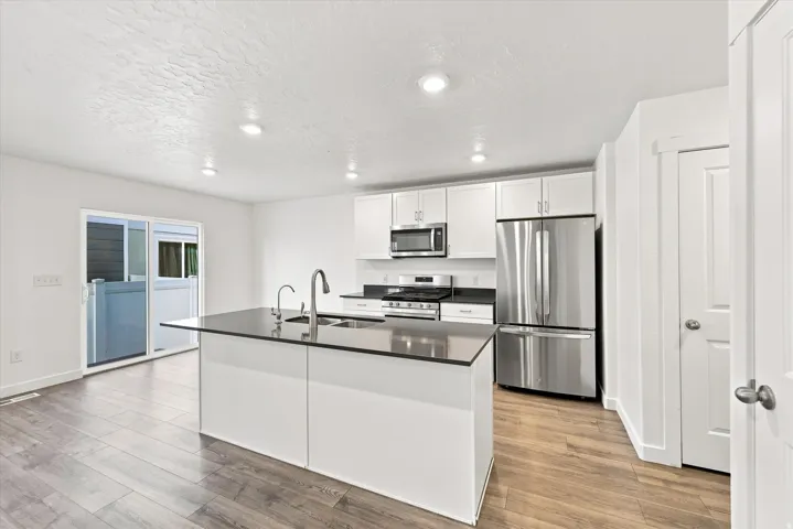 Kitchen featuring a central island with dual sinks, stainless steel appliances, white cabinetry, dark countertops, and wood-finish flooring