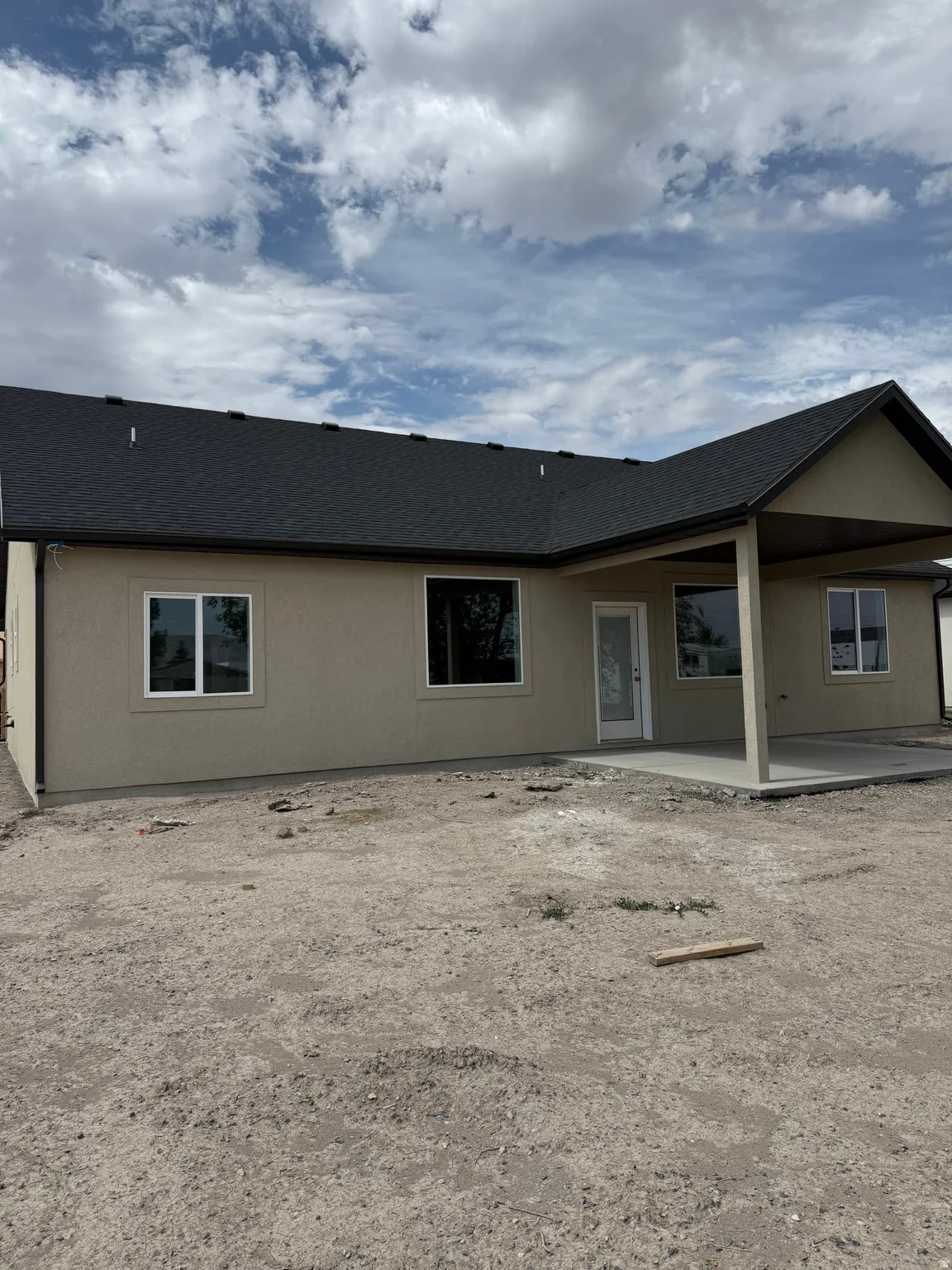 Single-story exterior with a tan stucco finish, dark shingle roof, white frame windows, and a covered patio with concrete flooring
