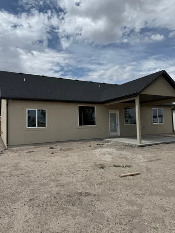Single-story exterior with a tan stucco finish, dark shingle roof, white frame windows, and a covered patio with concrete flooring