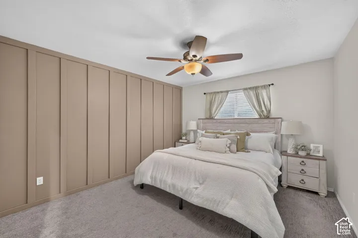 Accent wall with vertical paneling, light gray carpeting, ceiling fan with integrated lighting, window with blinds, and a distressed wood-finish headboard