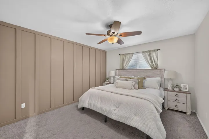 Accent wall with vertical paneling, light gray carpeting, ceiling fan with integrated lighting, window with blinds, and a distressed wood-finish headboard