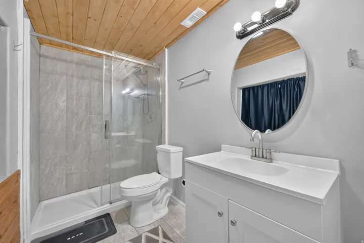 Wood-paneled ceiling, glass-enclosed shower, tile-surround shower, white vanity with integrated sink, and oval mirror