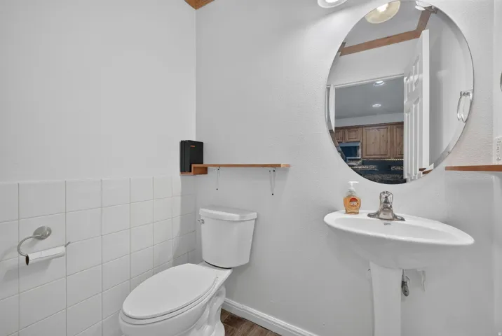 White pedestal sink with a round mirror, white tile wainscoting, and wood-finish flooring