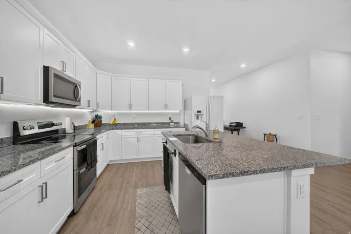 Open-concept kitchen featuring white cabinetry, granite countertops, stainless steel appliances, a kitchen island with sink, and wood-finish flooring
