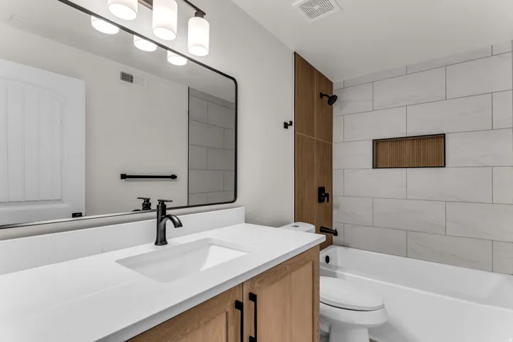 Modern bathroom featuring a single vanity with a white countertop and integrated sink, matte black fixtures, a rectangular framed mirror, and a tiled shower-bath combination with a built-in niche