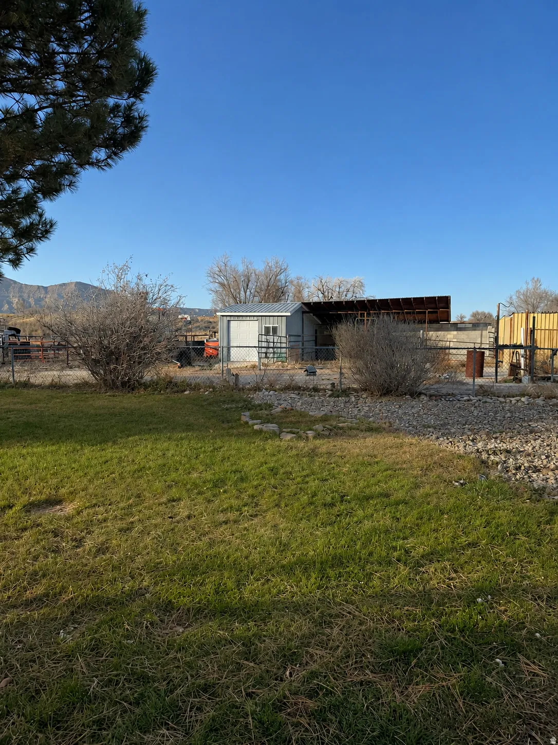 Shed with a metal roof and two windows, enclosed by a chain-link fence, with mountains in the background