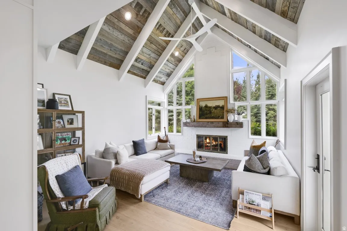 Vaulted ceiling with exposed wood beams, white brick fireplace with a wood-finish mantel, and large picture windows