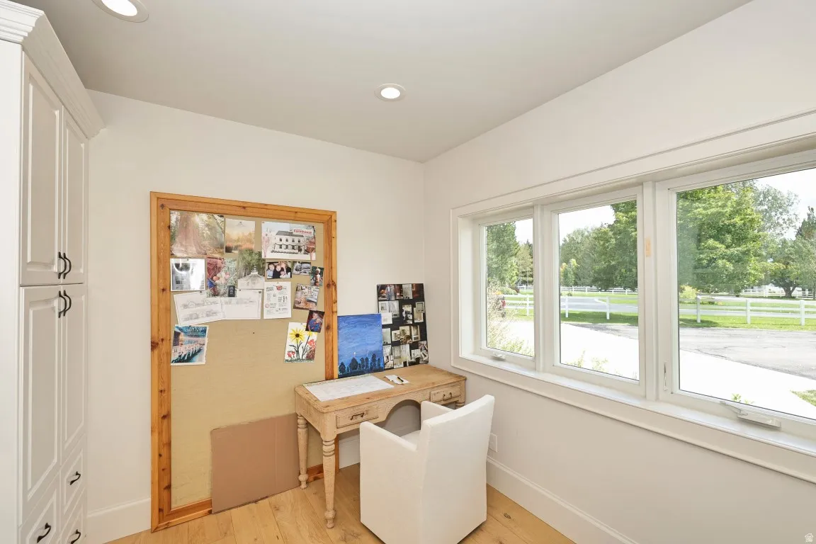 Bright home office featuring expansive multi-pane windows with natural light, warm wood-finish flooring, cork bulletin board with wooden frame, built-in desk with storage drawer, and white cabinetry