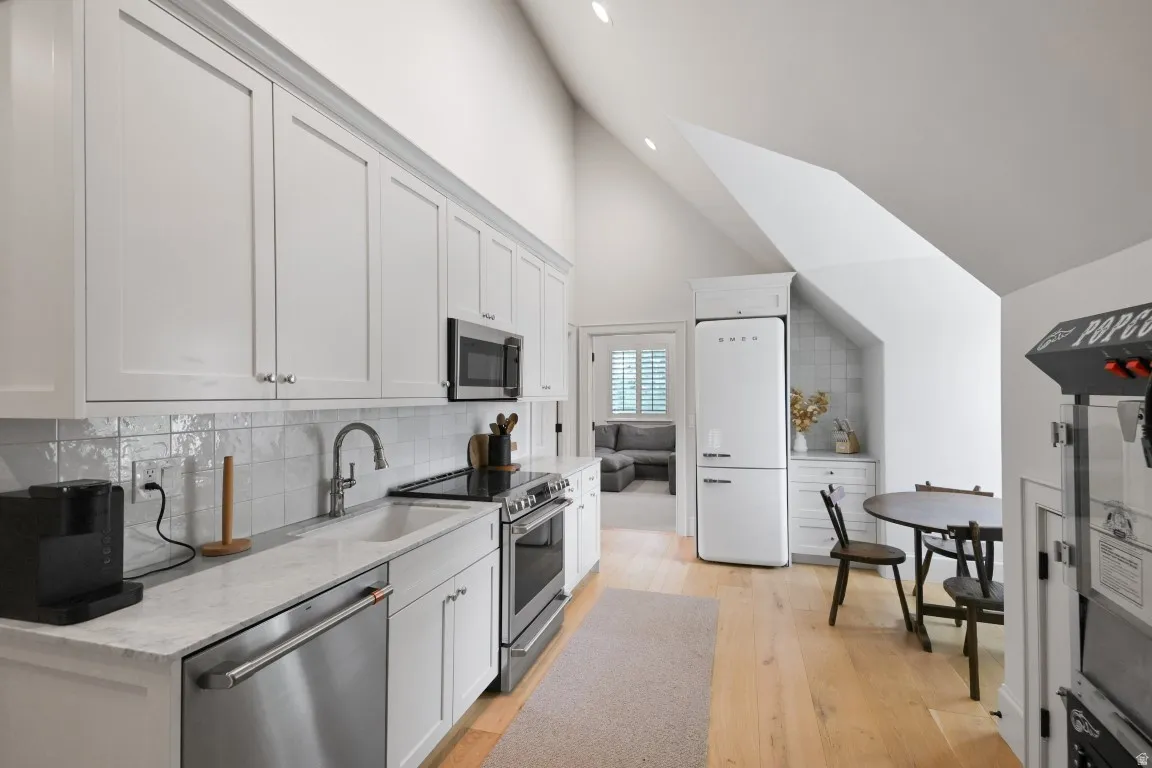 Vaulted ceiling kitchen featuring white shaker cabinetry, stainless steel appliances, a white farmhouse sink, and light wood-finish flooring
