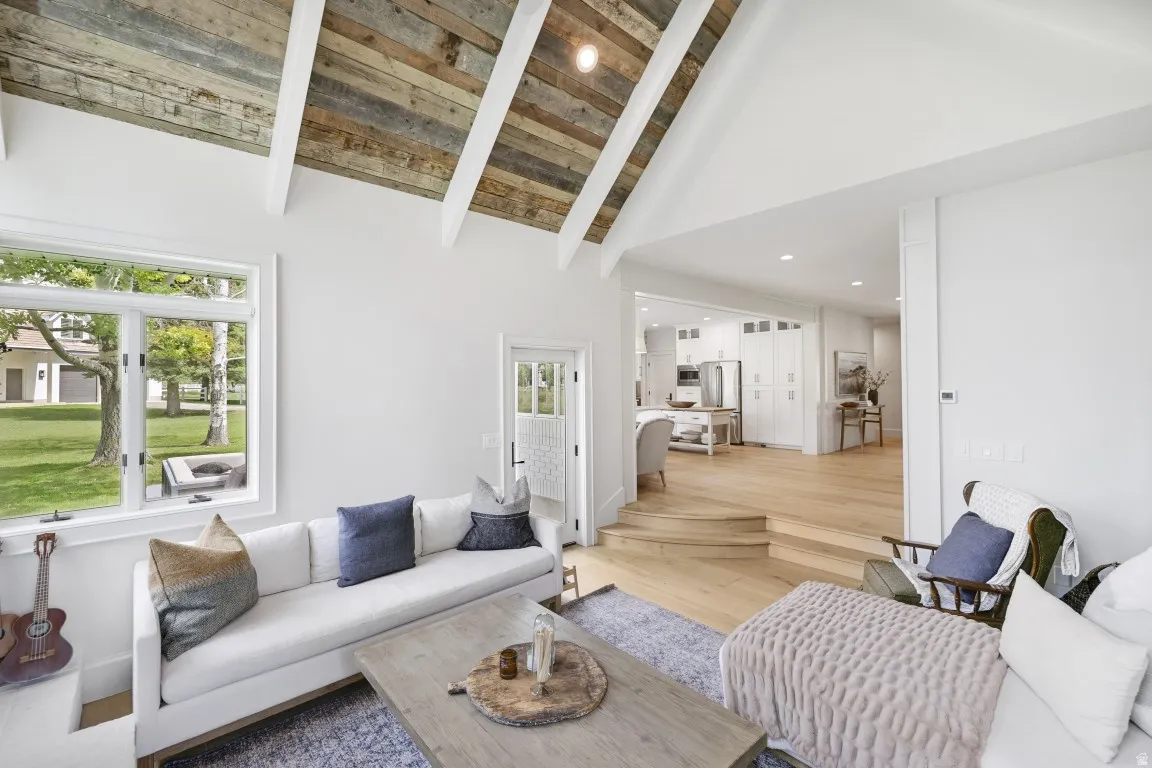 Vaulted ceiling with exposed wood beams, large windows, light wood-finish flooring, and crisp white walls