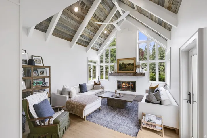 Vaulted ceiling with exposed wood beams, white brick fireplace with a wood-finish mantel, and large picture windows
