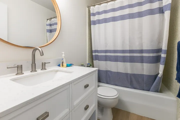 White vanity with recessed paneling and brushed nickel hardware, integrated sink with a light-toned countertop, brushed nickel widespread faucet, and a round mirror with a wood-finish frame