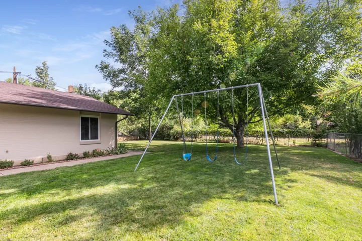 Expansive grassy lawn with a mature shade tree, chain link fencing, a paved pathway, and exterior siding