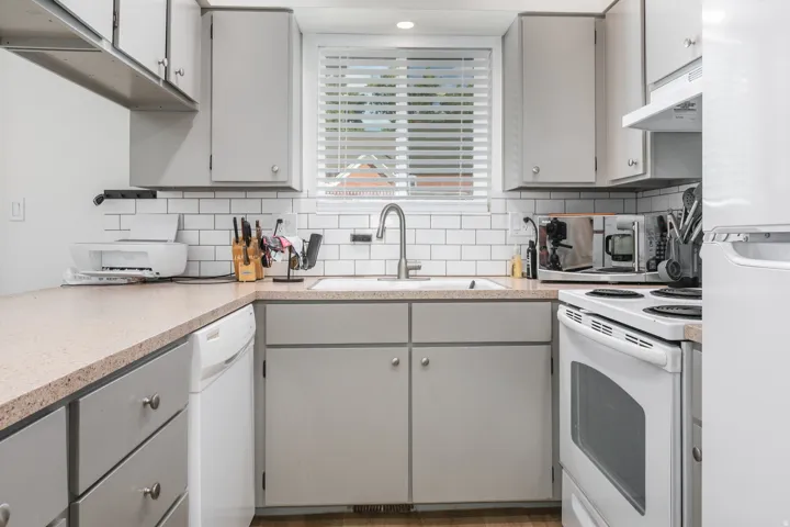 Kitchen featuring gray cabinetry, white subway tile backsplash, light-toned countertops, a stainless steel sink with a gooseneck faucet, and white appliances