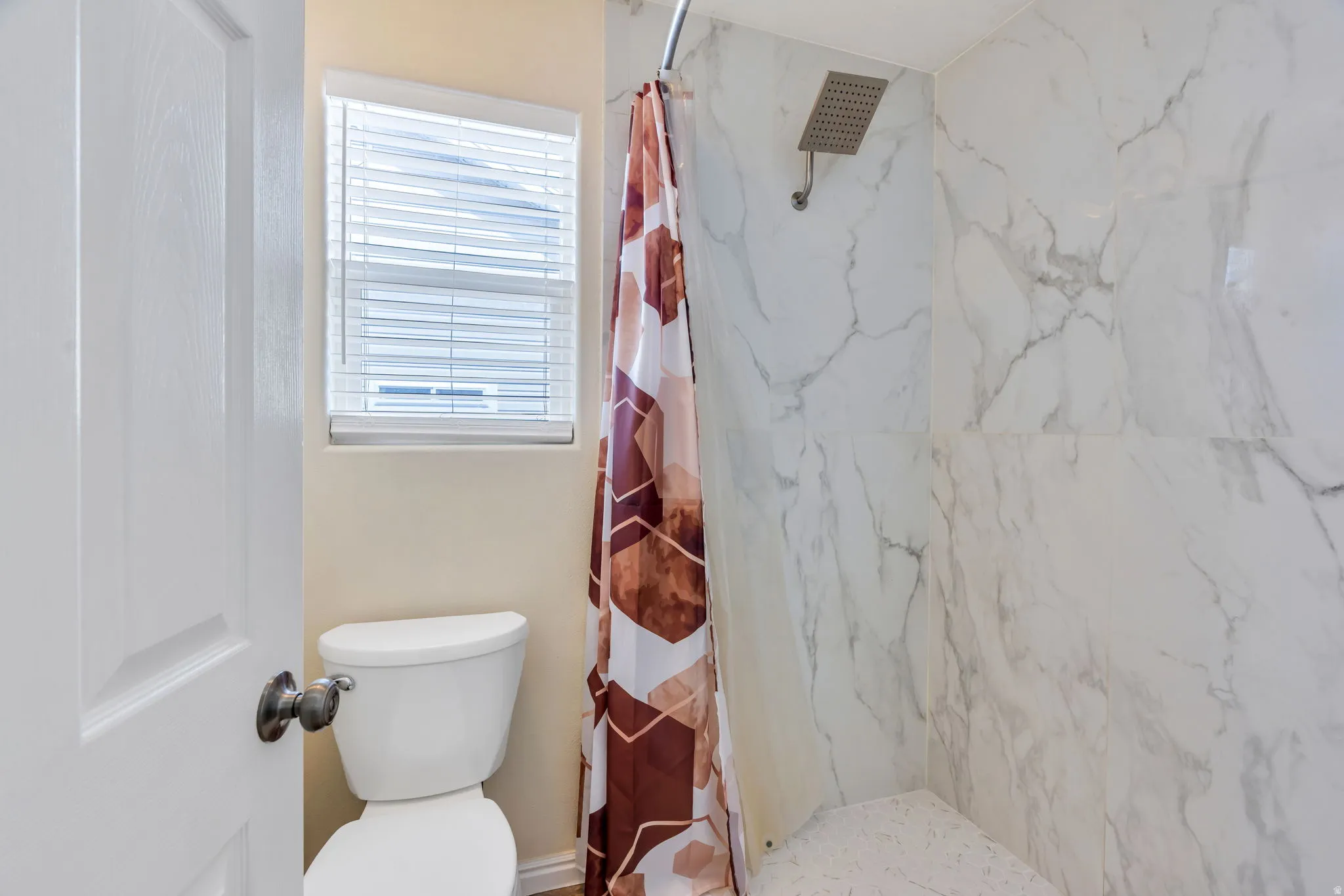 White toilet with a window above, white shower with patterned wall tile, and a rain shower head