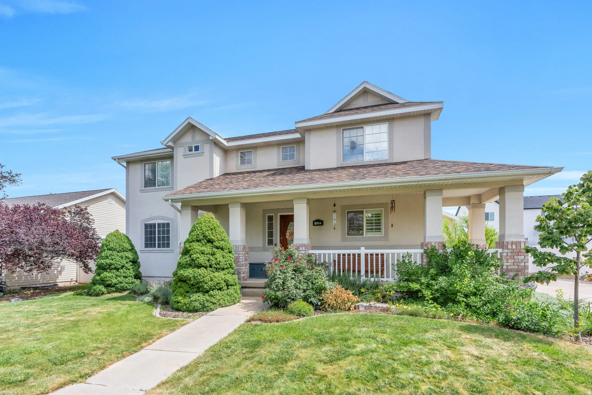 Two-story stucco exterior with dormer windows, covered front porch with white railing, stone column accents, and a concrete walkway