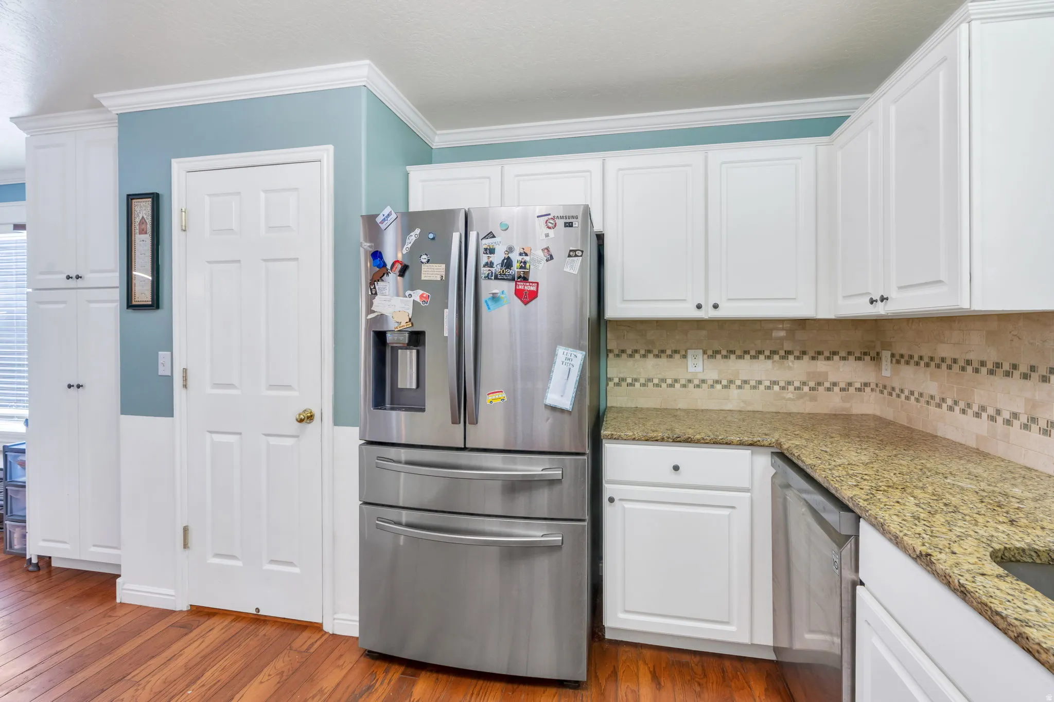 White cabinetry and crown molding, stainless steel refrigerator, granite countertops, tile backsplash, and wood-finish flooring