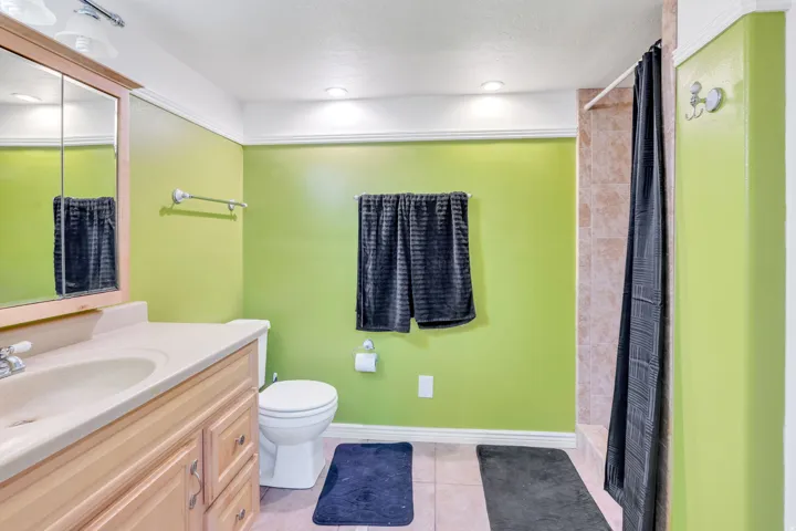 Recessed ceiling lighting, wood-finish vanity with countertop sink, wall-mounted mirrored medicine cabinet, tile flooring, and a tiled shower surround