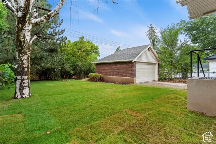 Expansive lawn area featuring a mature birch tree, a detached brick garage with a gabled roof, a concrete driveway, and established perimeter landscaping