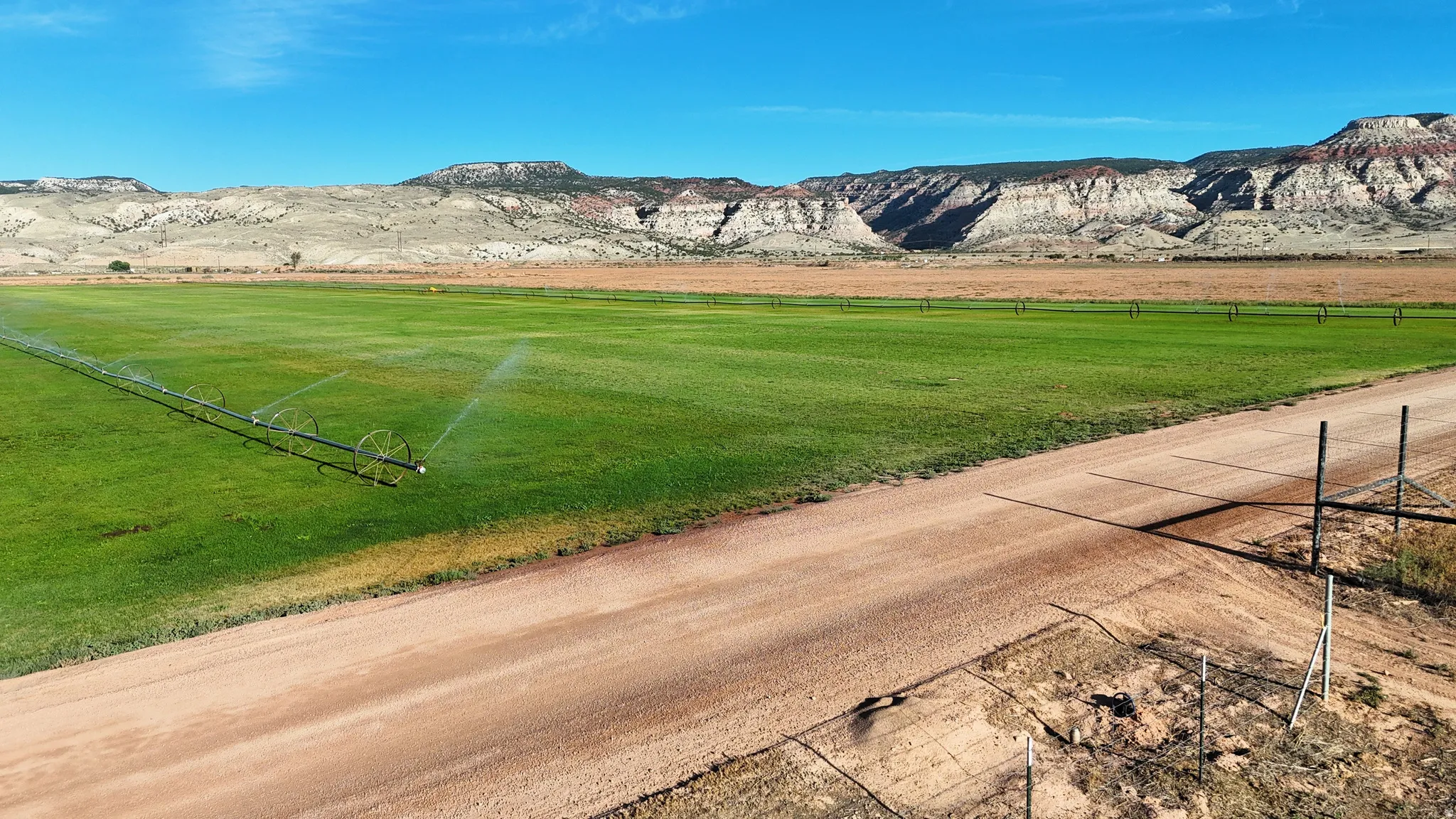 Expansive green field featuring a pivot irrigation system, a natural dirt road, and distant rugged mountains