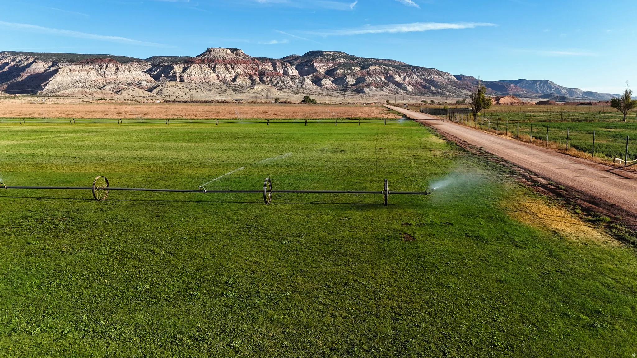 Expansive green fields with an irrigation system, unpaved road, and distant mountain backdrop