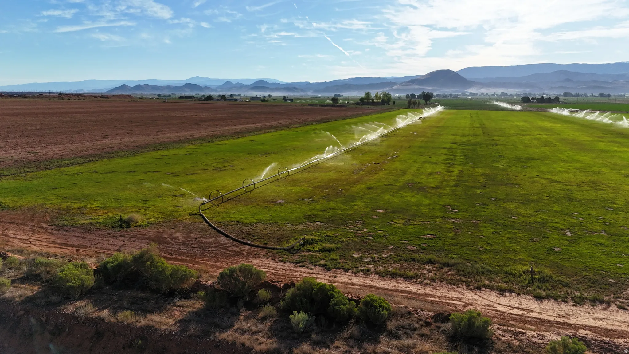 Expansive agricultural land featuring active irrigation systems across verdant fields and tilled acreage, with distant mountainous terrain