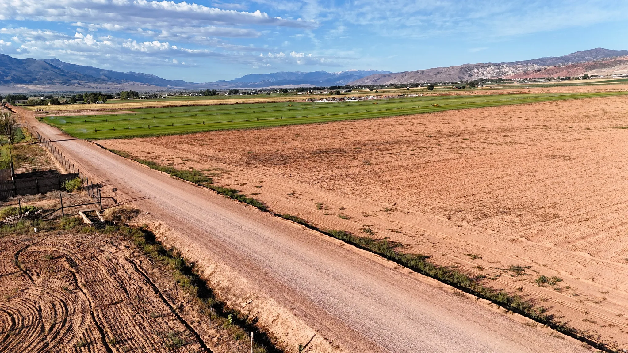 Expansive land featuring a gravel road, tilled fields, green agricultural areas, and distant mountains