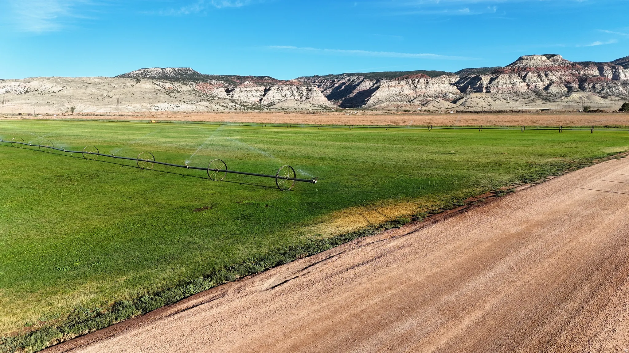 Expansive green pasture with irrigation systems, bordered by a dirt road and distant mountains