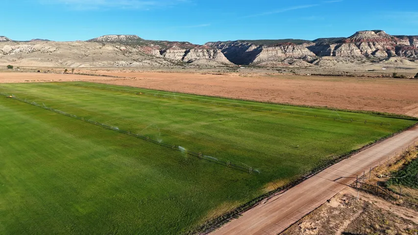 Expansive green field with irrigation system, bordered by a dirt road and fencing, set against a backdrop of distant mountains