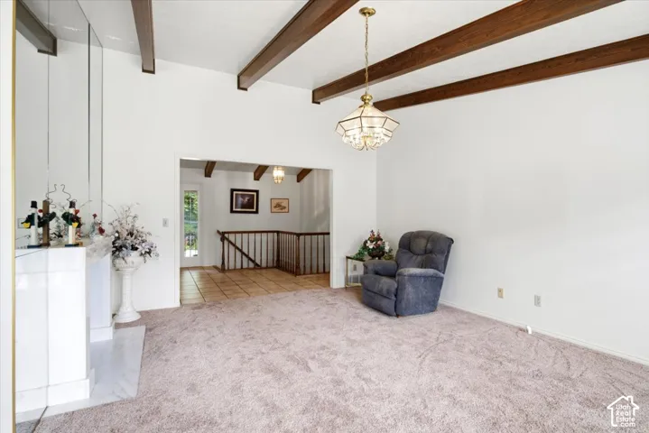 Vaulted ceiling with exposed wood beams, overhead chandelier, white fireplace surround with reflective paneling, and tile flooring visible in the entry