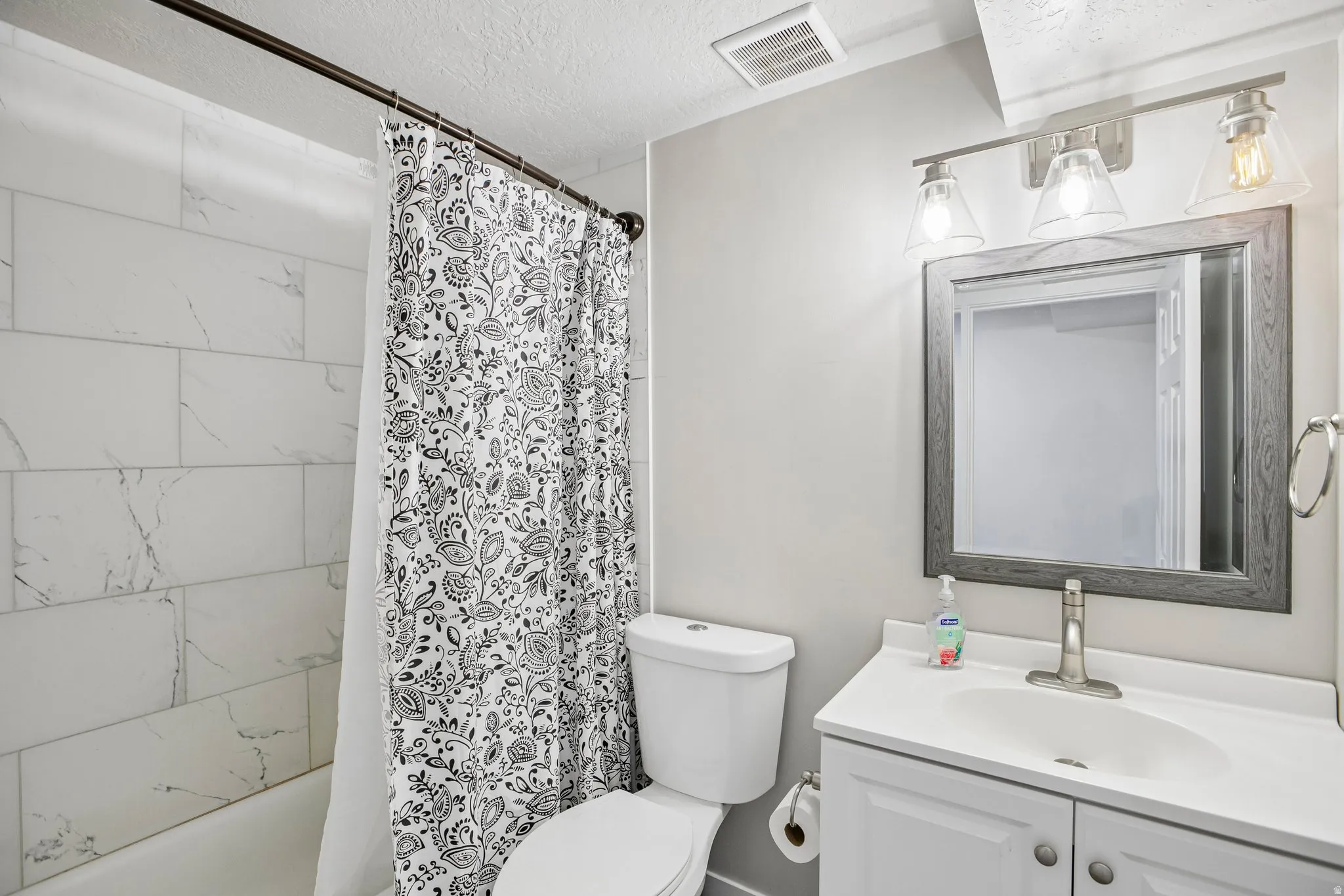 Bathroom featuring a white vanity with integrated sink, brushed nickel faucet, framed mirror, three-light vanity fixture, and tiled shower surround