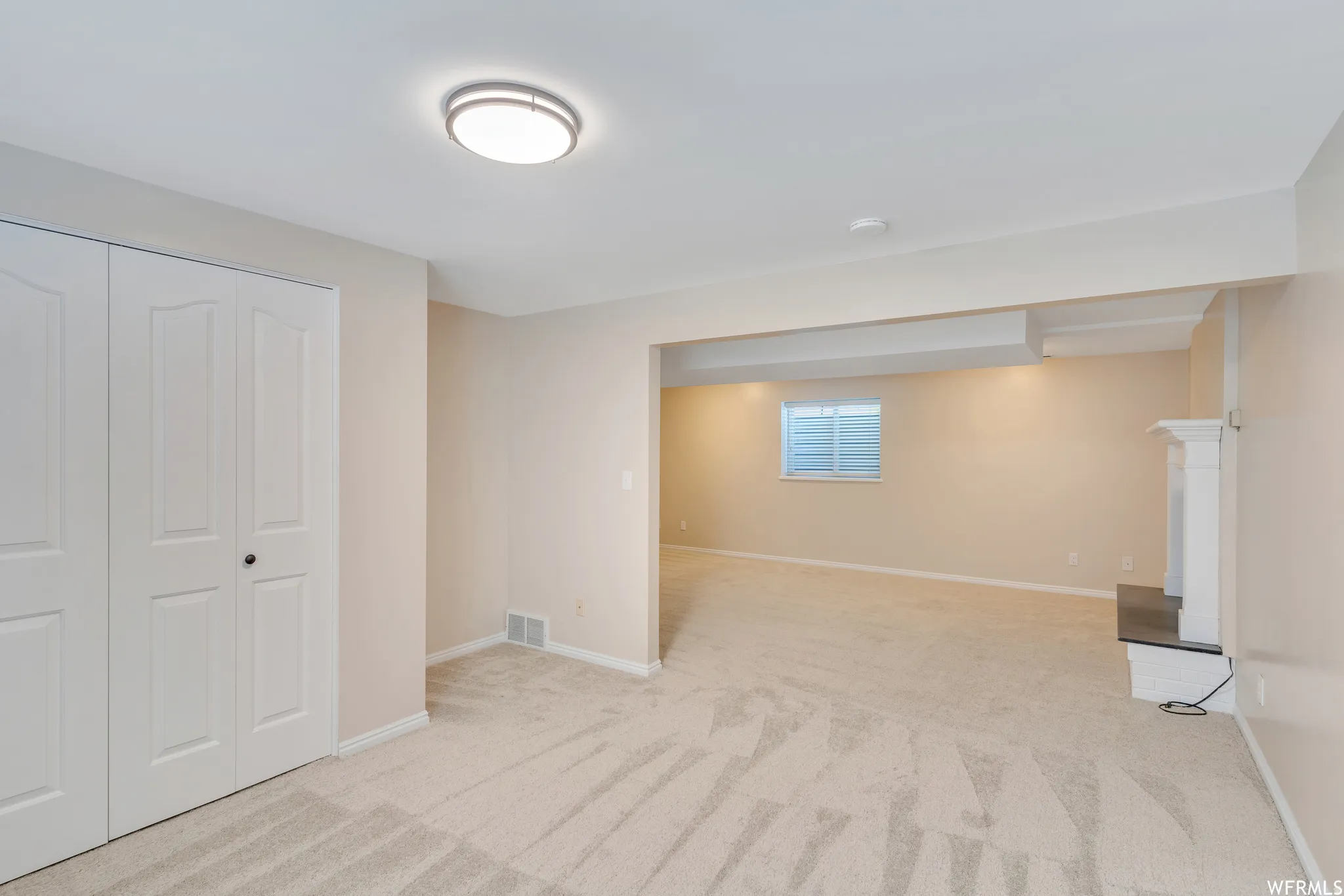 Neutral tone wall paint, light-colored carpeting, white bi-fold closet doors, white mantel with dark hearth, and a single window with blinds